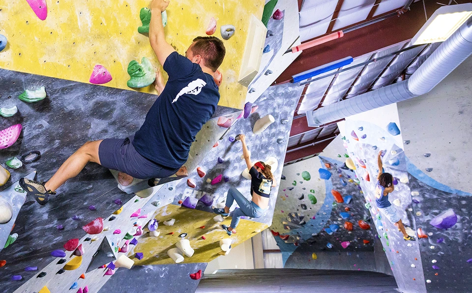A man is bouldering over the lip of an overhanging wall. In the background a woman is also bouldering.