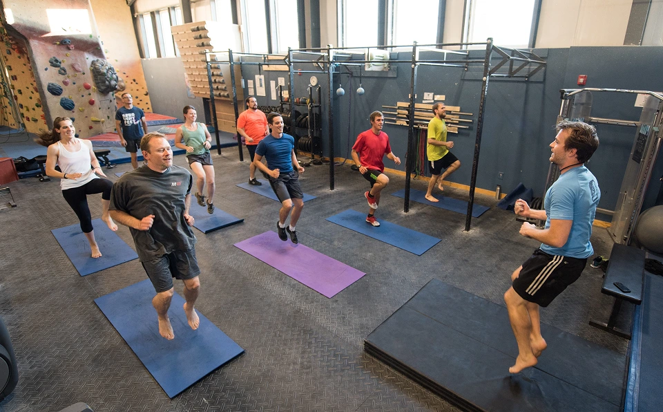 An instructor guides a group fitness class. Each person is jumping on a yoga mat next to a fitness space and a bouldering wall.