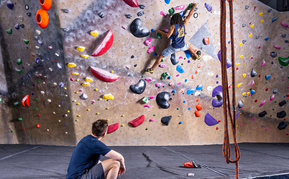 A man with dreads in a tank top is reaching up to the next hold on a boulder problem. A man sits on the edge of the mat and watches.