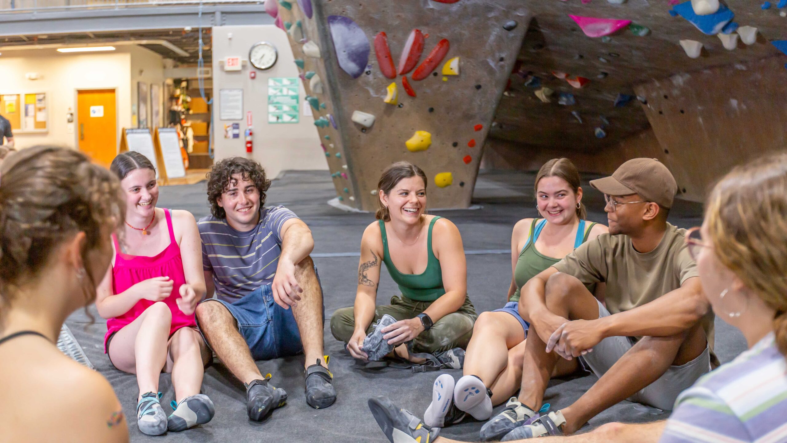 A group of people sit together on a bouldering mat in front of the wall laughing and talking together.