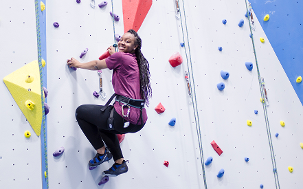 A smiling person with long hair climbs an indoor rock wall in a maroon shirt and harness. The wall features colorful holds on a white and blue surface.