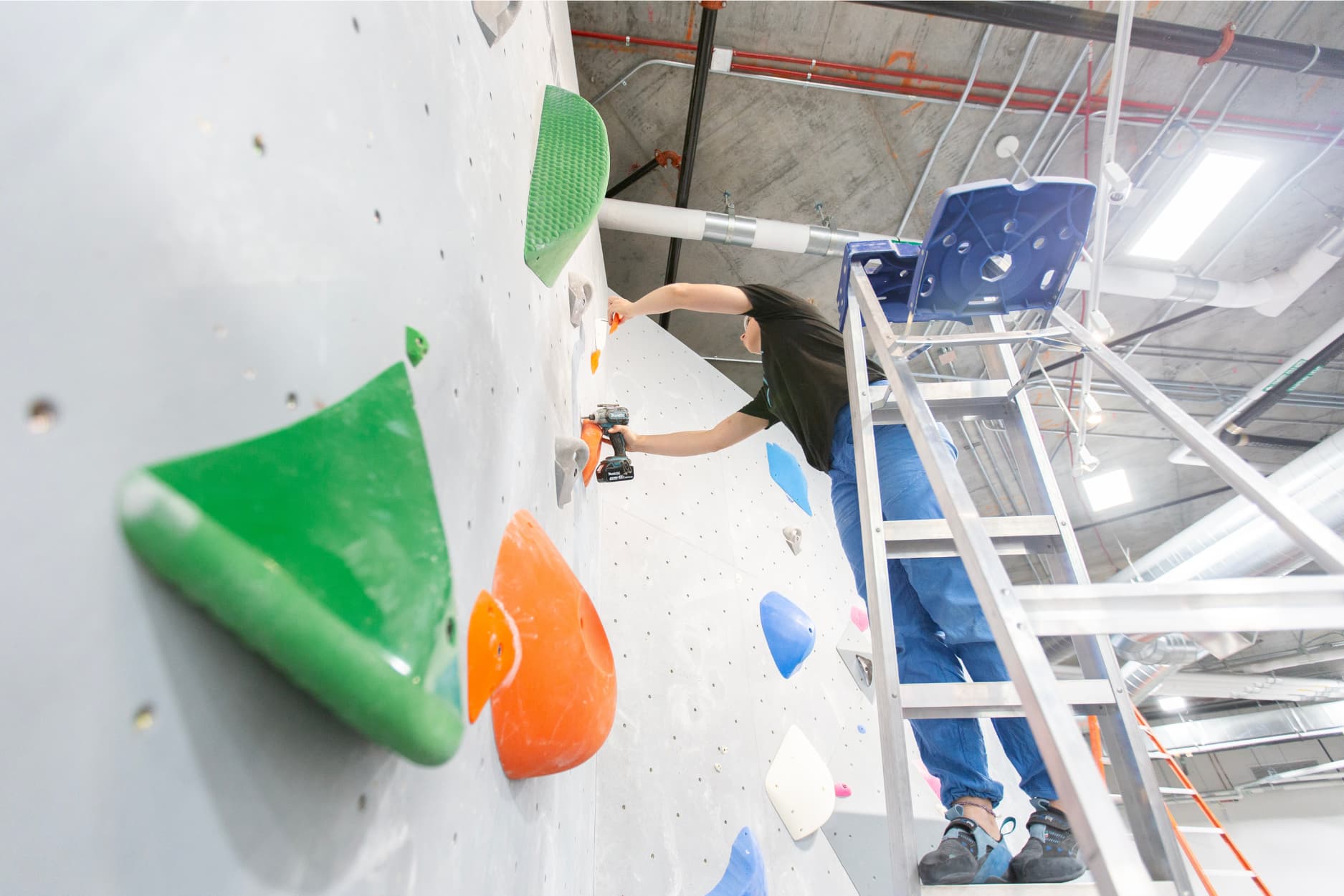 A woman stands on a ladder as she drills a hold into a climbing wall.