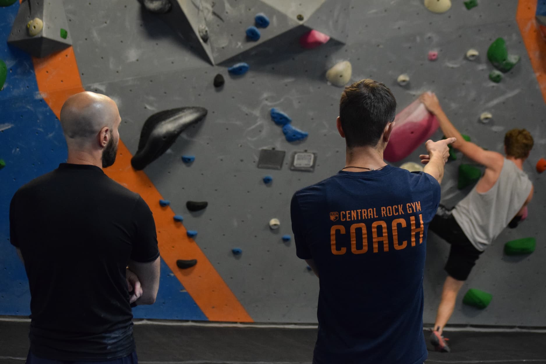 A Man wearing a COACH shirt gives direction to a man on the bouldering wall while a bald man watches.