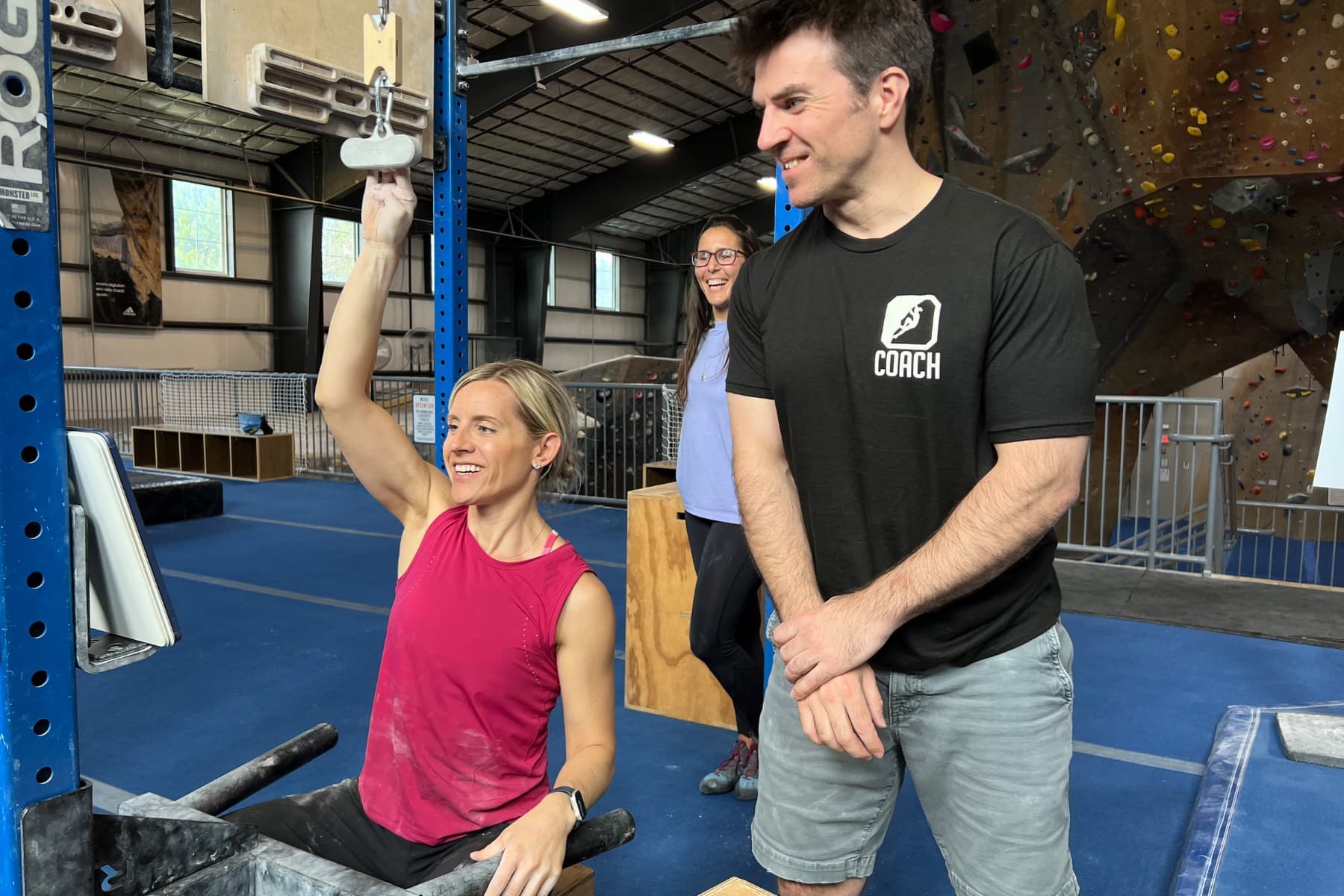 A Central Rock Gym coach stands by a woman who is training finger strength using a Tension block.