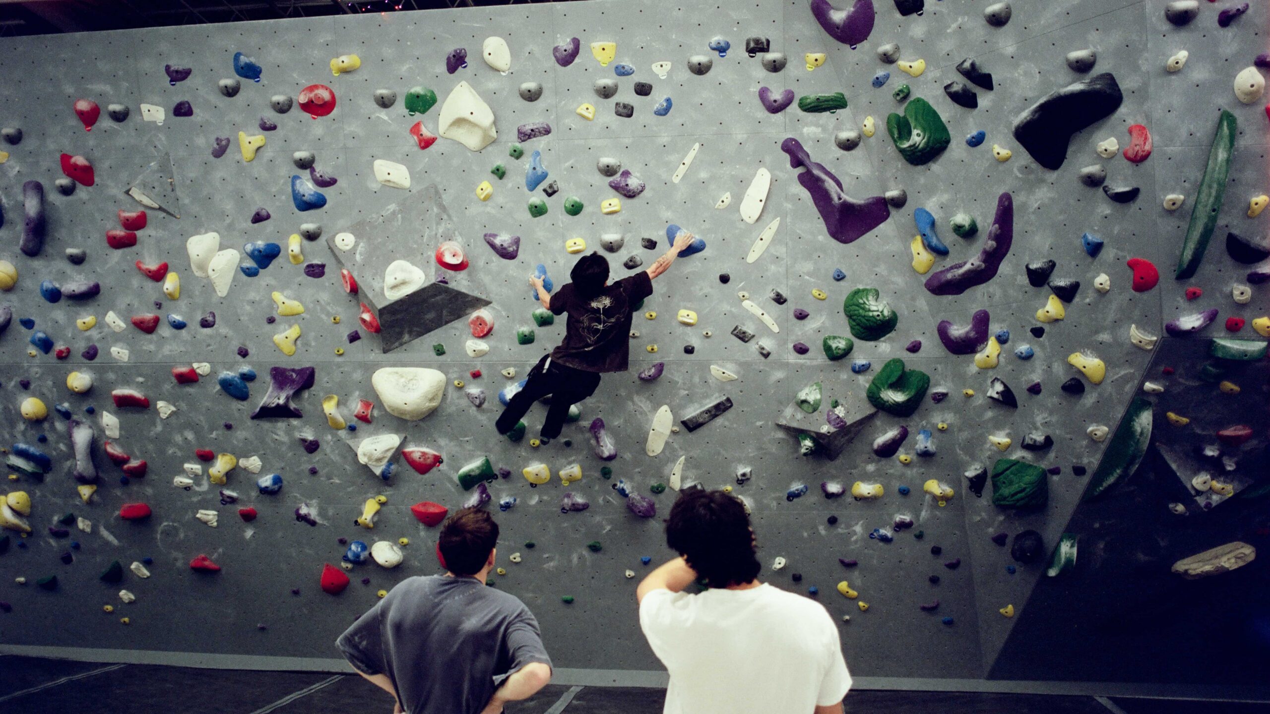 Two men watch another man bouldering on the wall during an adult team practice.