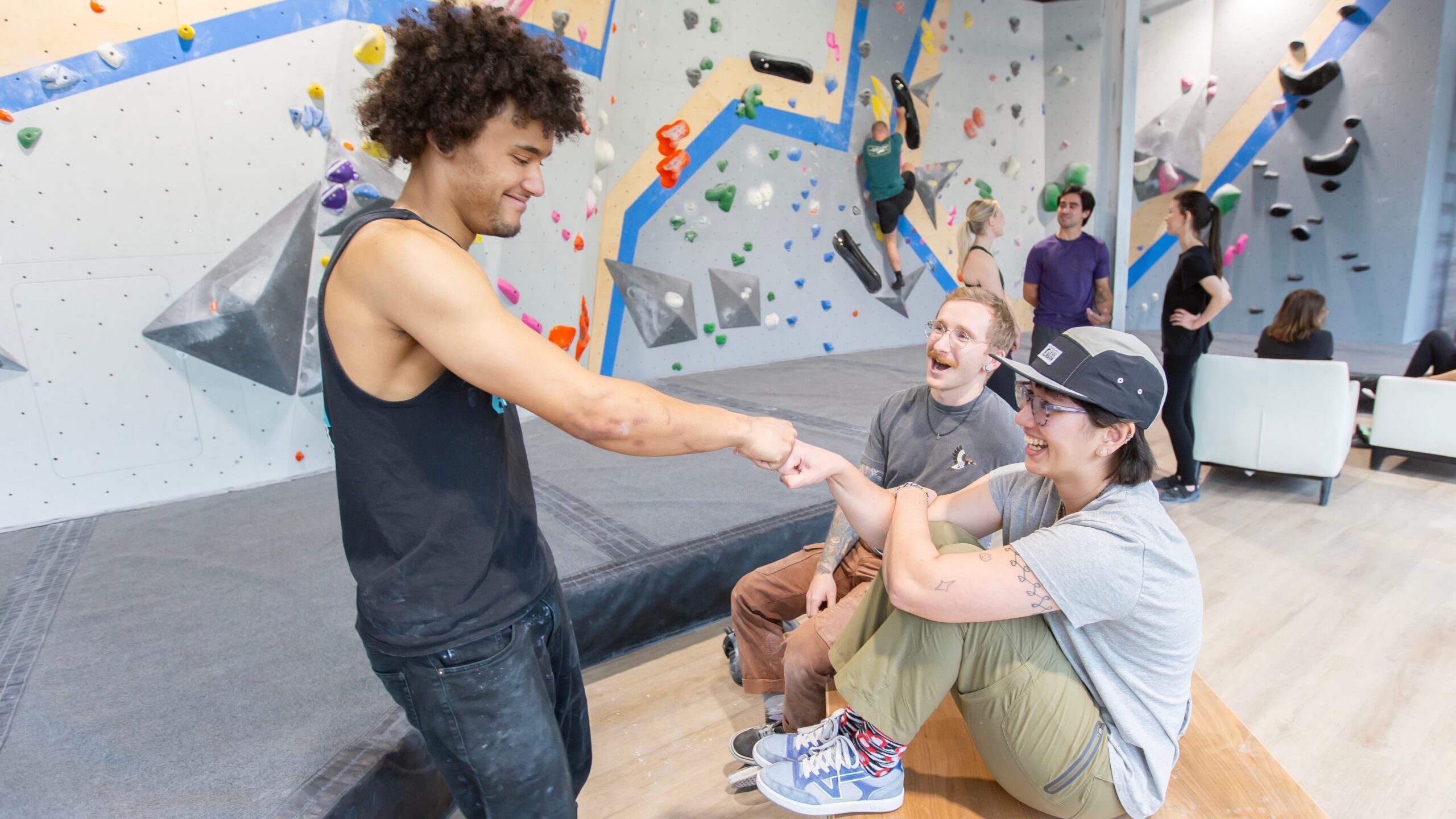 A man with an afro fist bumps his friend. She is sitting crouched on a bench and is wearing a hat. Next to her another friend cheers.