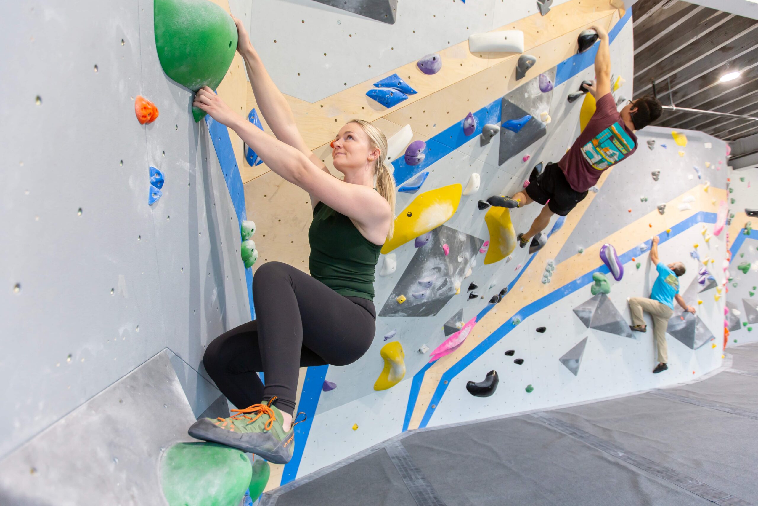 A woman with her hair in a ponytail crouches on the bouldering wall as she leans into a sidepull crimp. In the background two men are also bouldering.