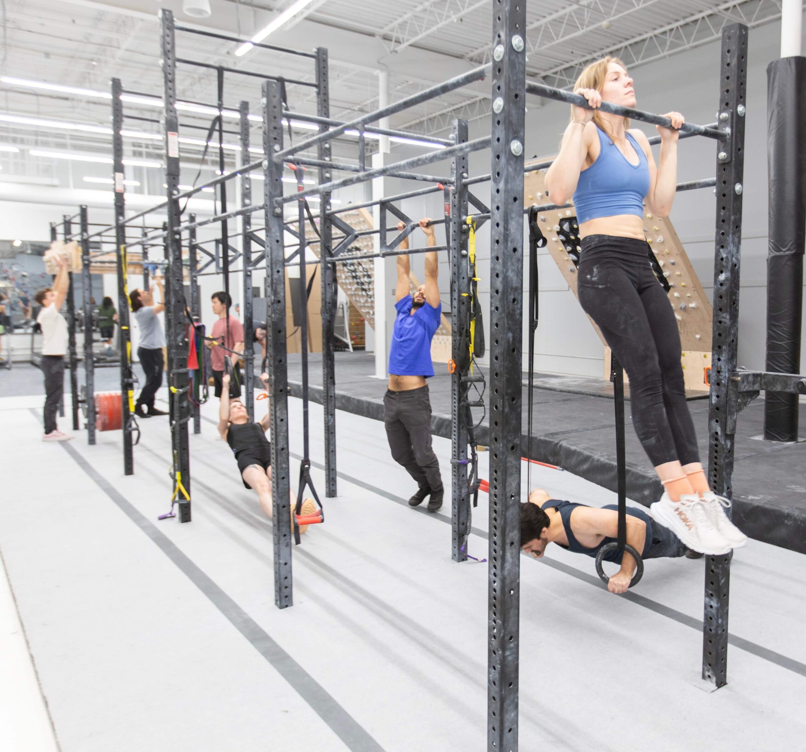 People are working out. In the foreground a woman is training pullups. Behind her other climbers are training with gymnastics rings and hang boards