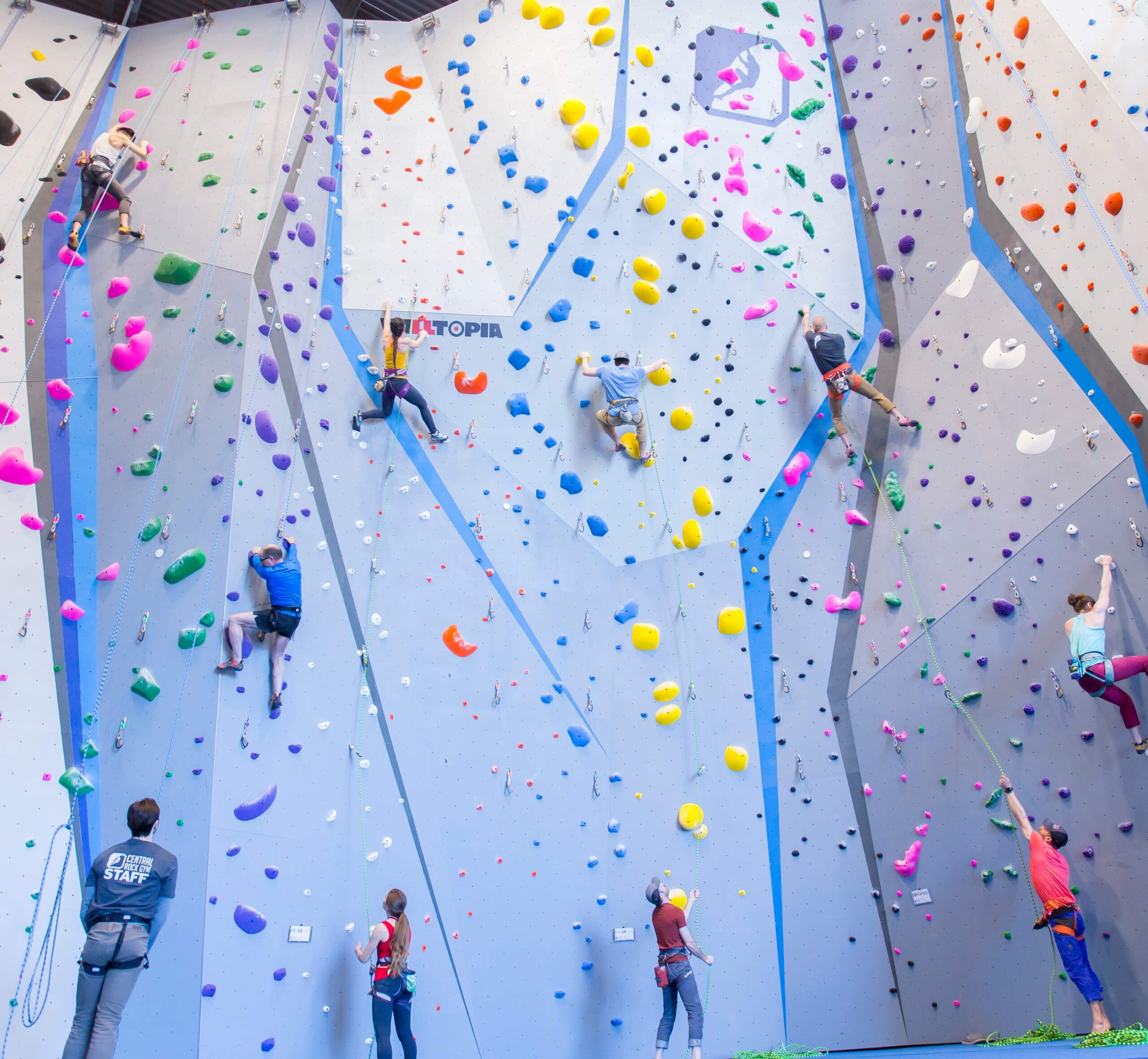 A wide shot of many pairs of people rope climbing on climbs of various difficulties. The wall has the CRG logo on it.