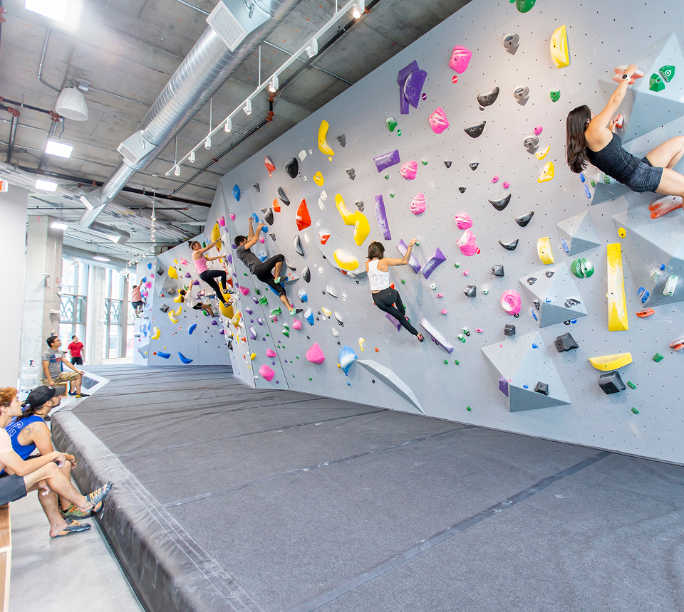People climb a vibrant indoor bouldering wall with multicolored holds. Others sit on padded floors. The scene conveys energy and focus.