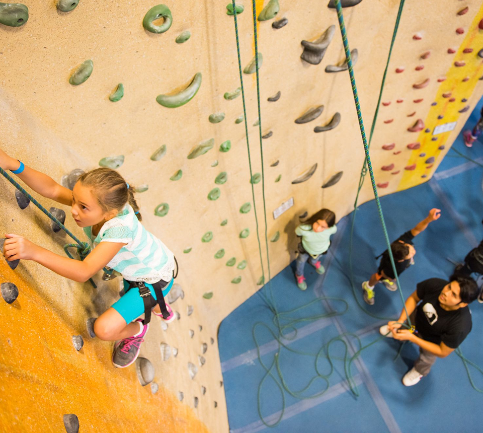 A young girl in a striped shirt climbs a colorful indoor rock wall with determination. Below, others watch and cheer, creating an atmosphere of encouragement.