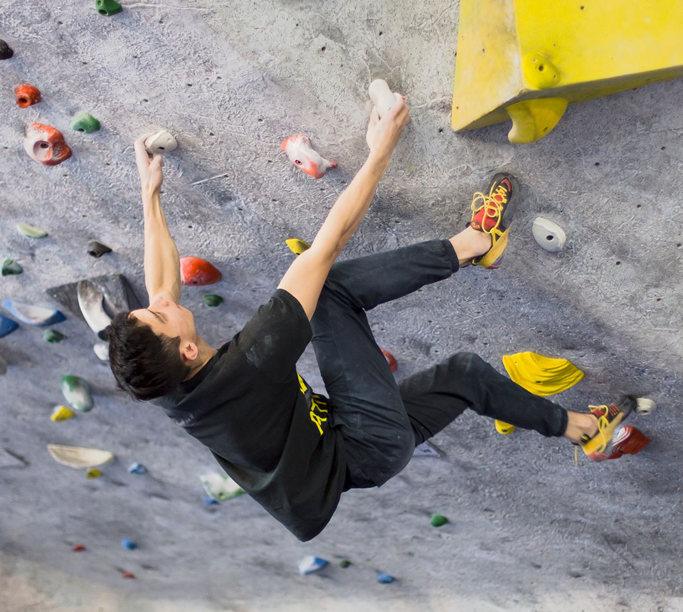 A man in a black shirt and climbing shoes tackles an indoor bouldering wall, gripping colorful holds with focus and determination.