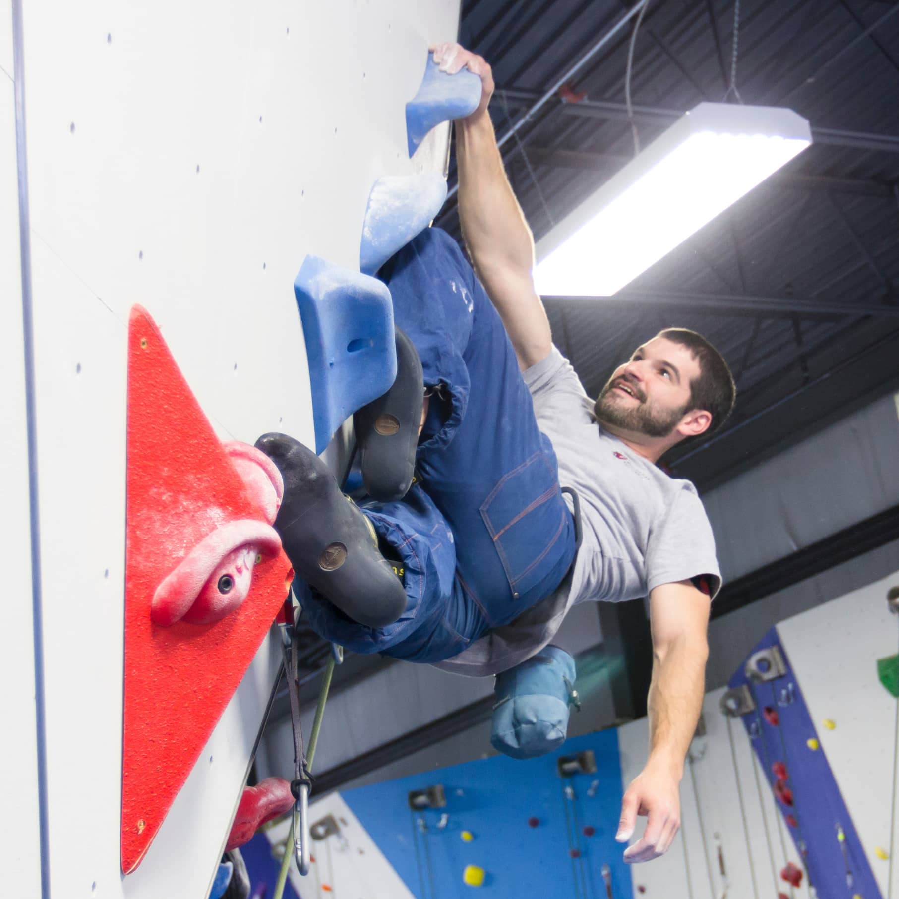 A man smiles as he lead climbs. He is backflagging with one foot as he rests on the climb and lets one arm hang.