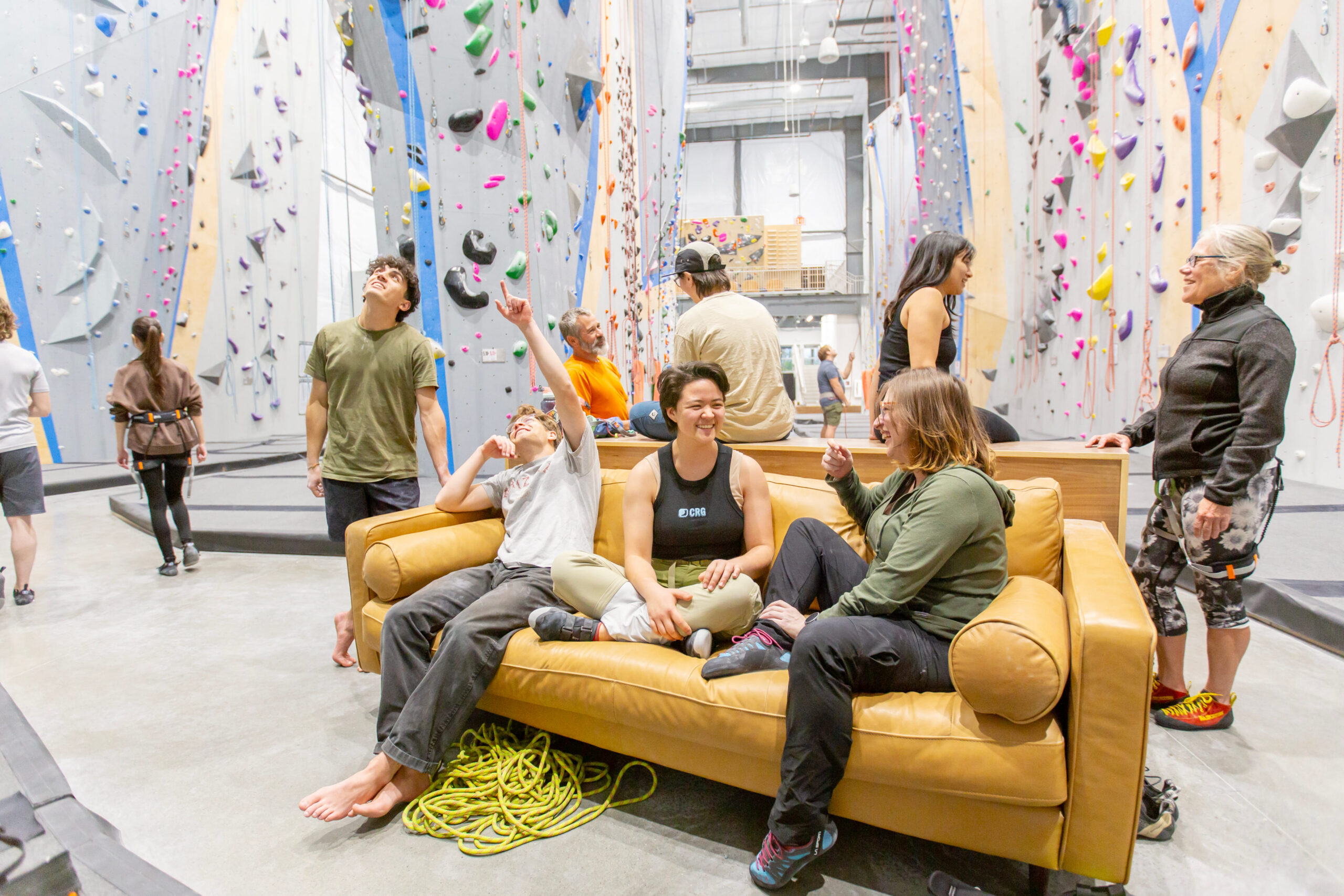 A group of people socializing on the couchg in front of the rope climbing walls. Two women sit and chat while one man points up at the wall, pointing out a climb to his friend.
