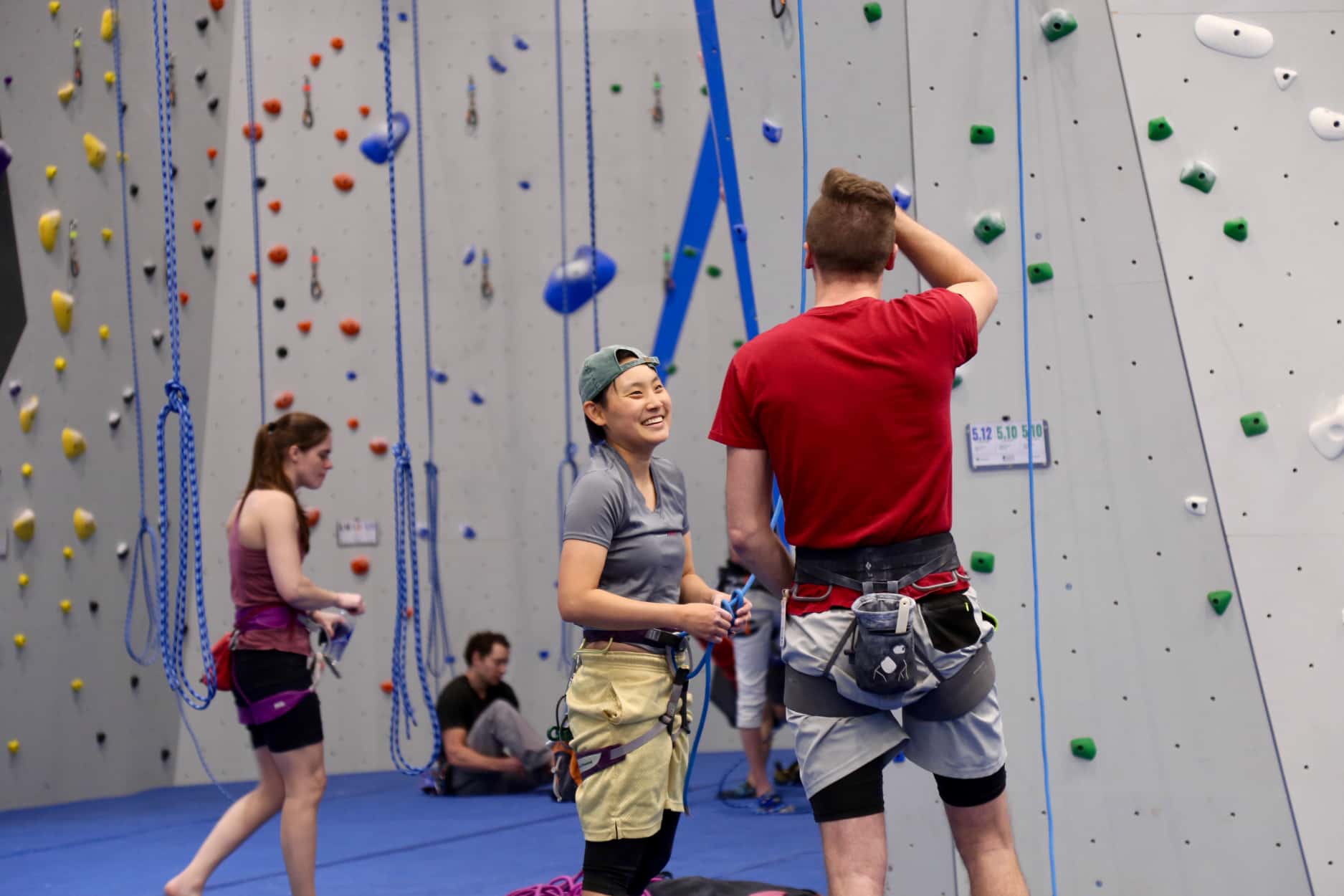 A tall man a short woman with a backwards cap rope climb together and laugh and talk together as they tie in for the belay.
