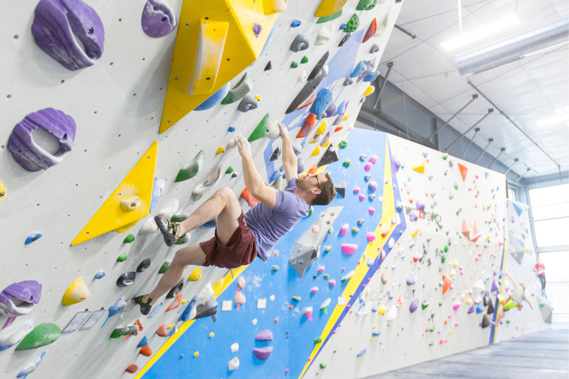 A bearded man steps his foot up a steep bouldering wall as he hangs with his arms stretched out.