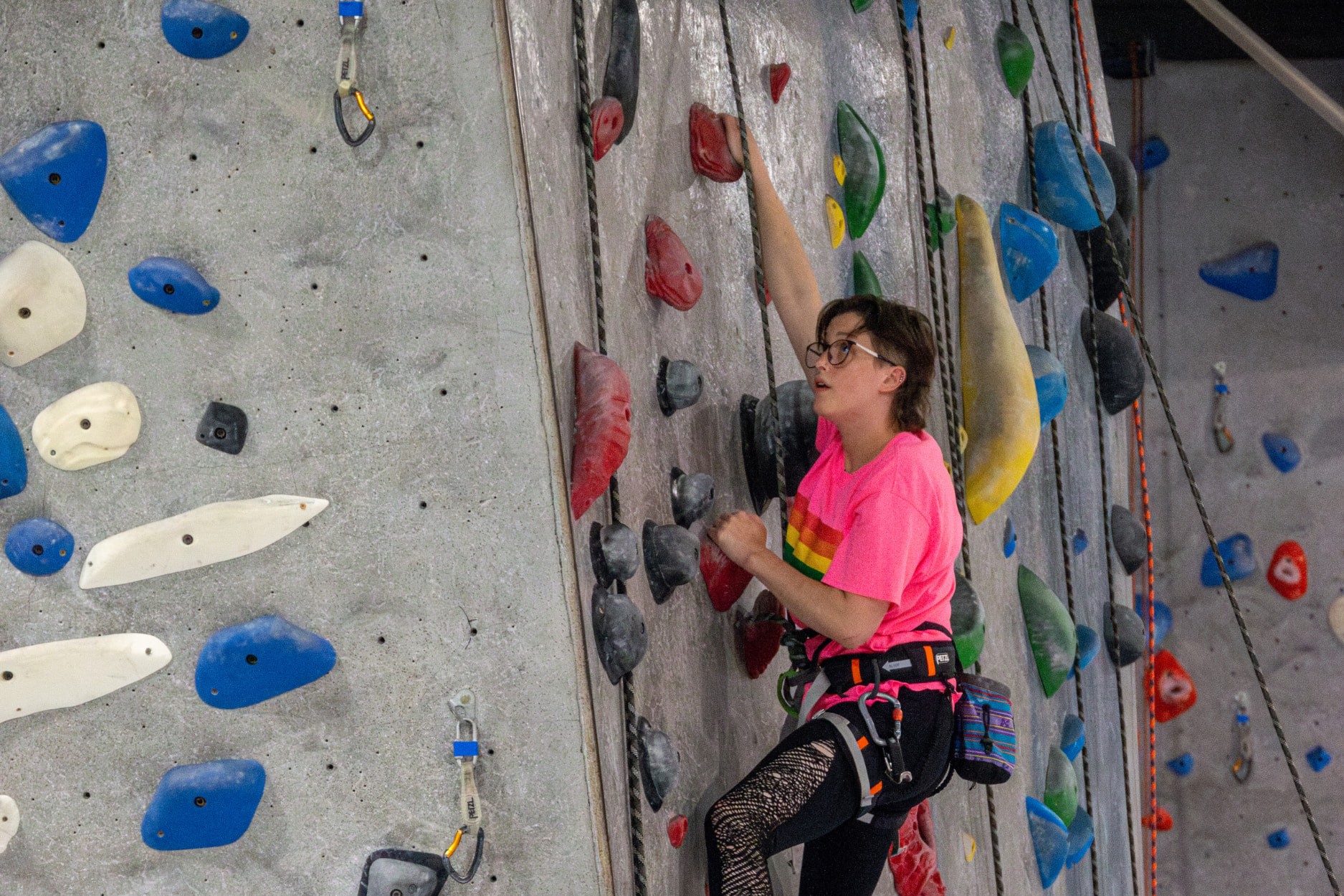 A woman wearing a pride tshirt top rope climbs on a slab wall. On her harness is a chalk bag and belay device.