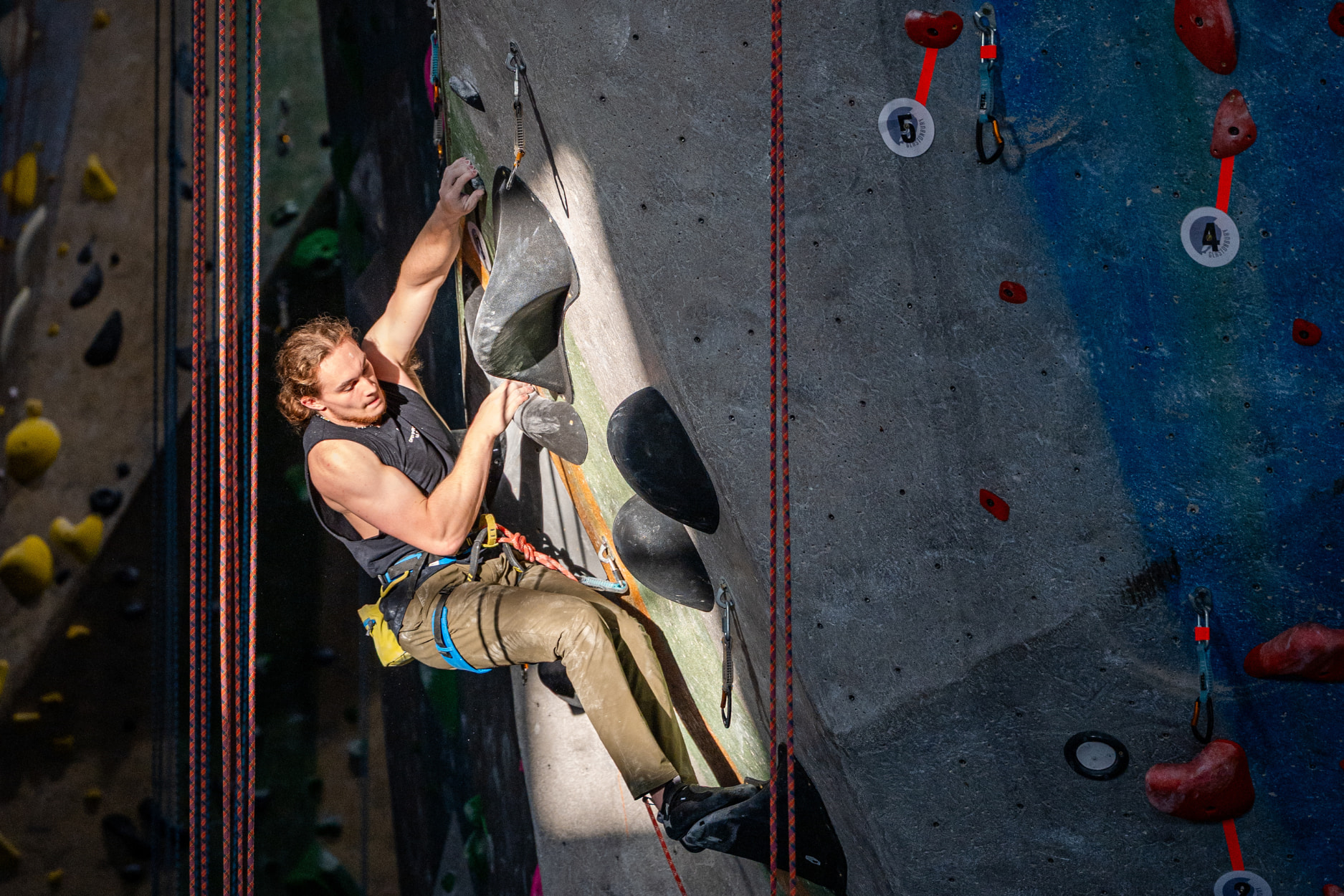 A man with long hair lead climbs through a patch of sunlight which is illuminating a rectangle on the lead climbing wall at Central Rock Gym.