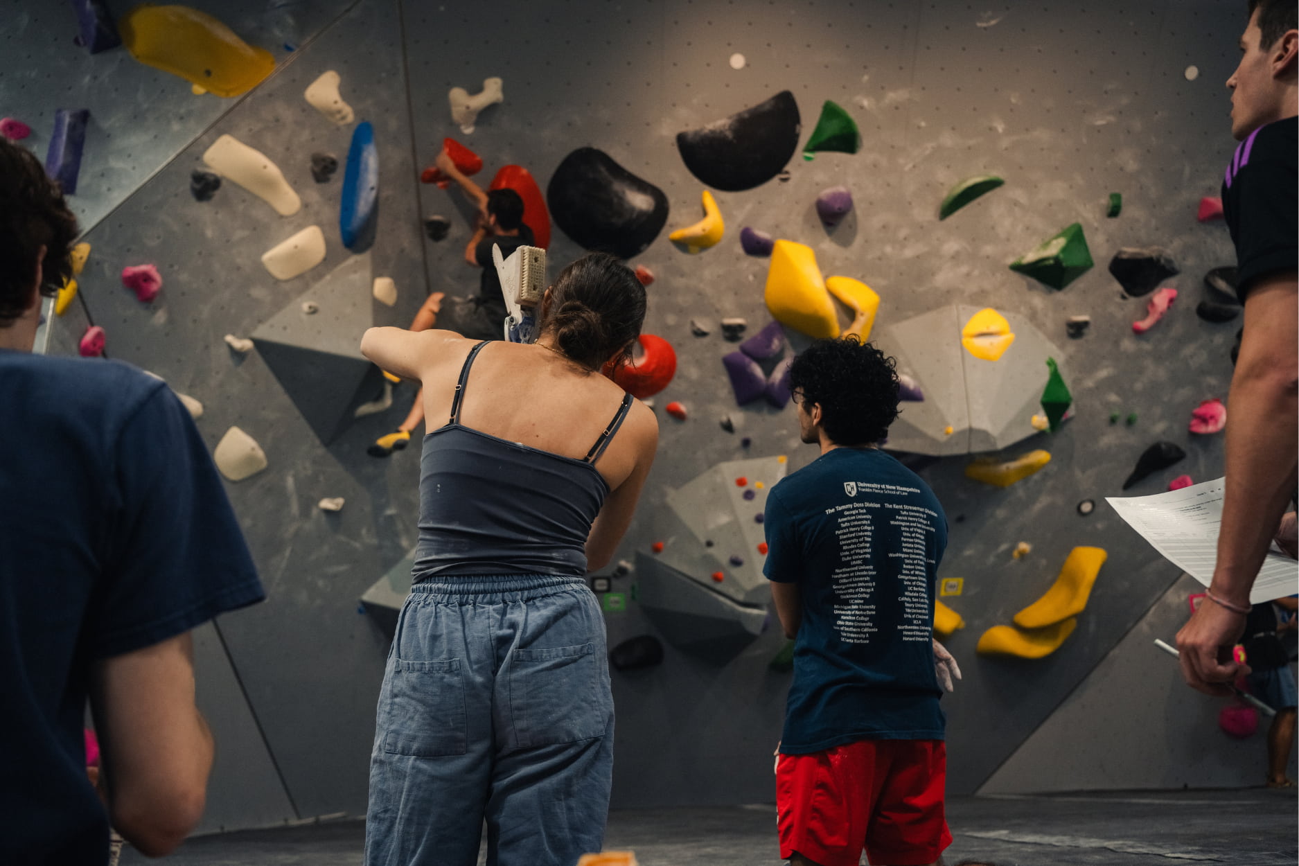 A woman stands looking at the bouldering wall, holding a extendable climbing brush and talking to a man about the beta of a boulder. Another woman is climbing on the wall.