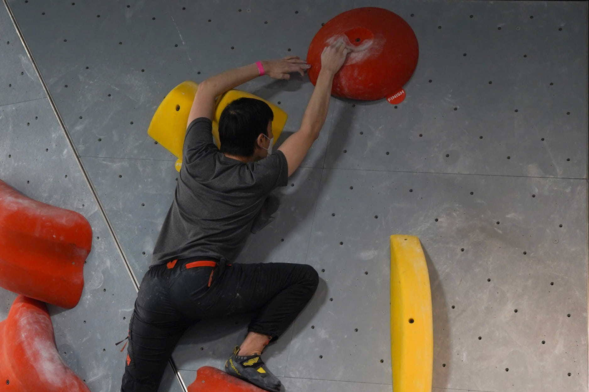 A man reaches up to the slopey finish hold of a climb and taps it with his second hand for the send. He is crouched low over his foot.