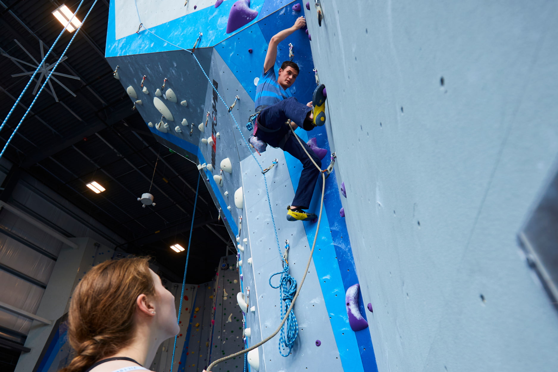 A woman with a braid belays for a man who is lead climbing.