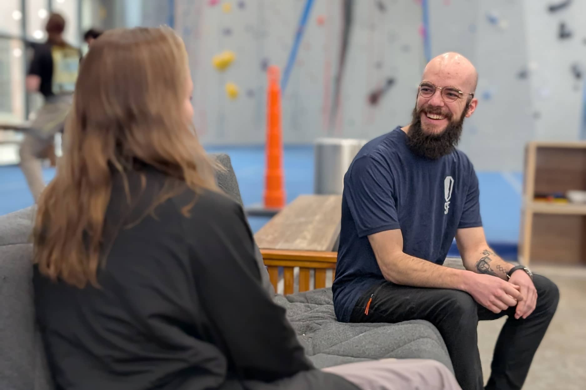 A Central Rock Gym instructor sits and answers the question from a member. He is bald and has a large beard. She has long wavy hair.