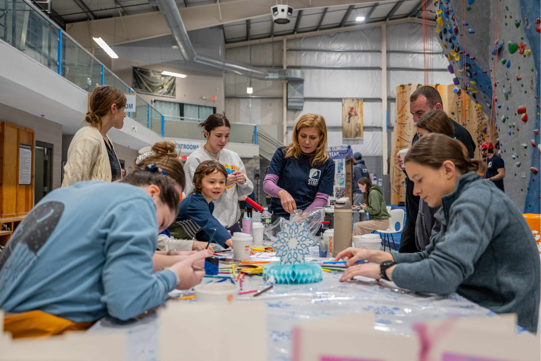 A group of men, women, and children gather around a winter crafts table at an event at Central Rock Gym. A CRG staff member provides guidance as she oversees the table and helps the guests with their crafts.