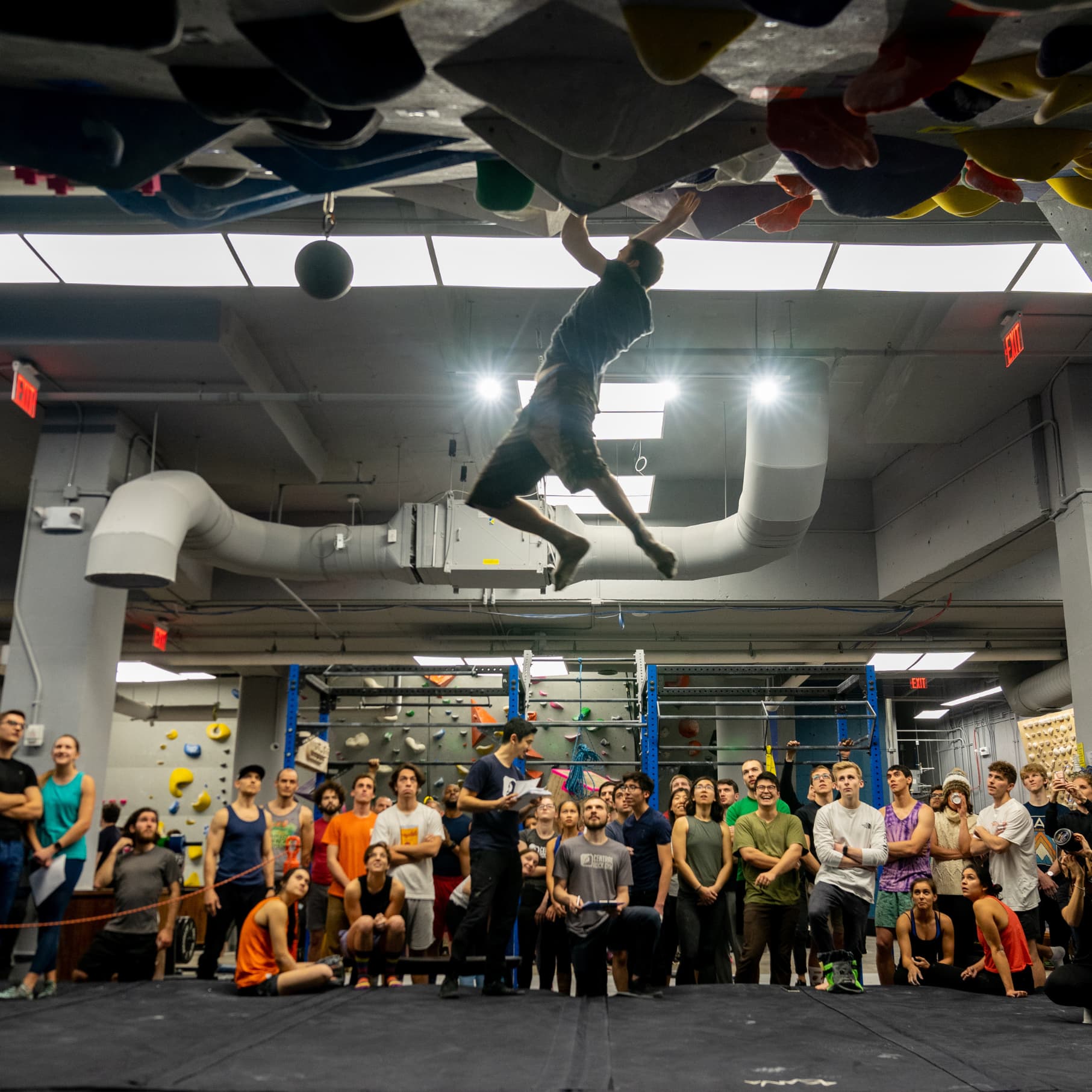 A man bouldering while many people spectate at central rock gym