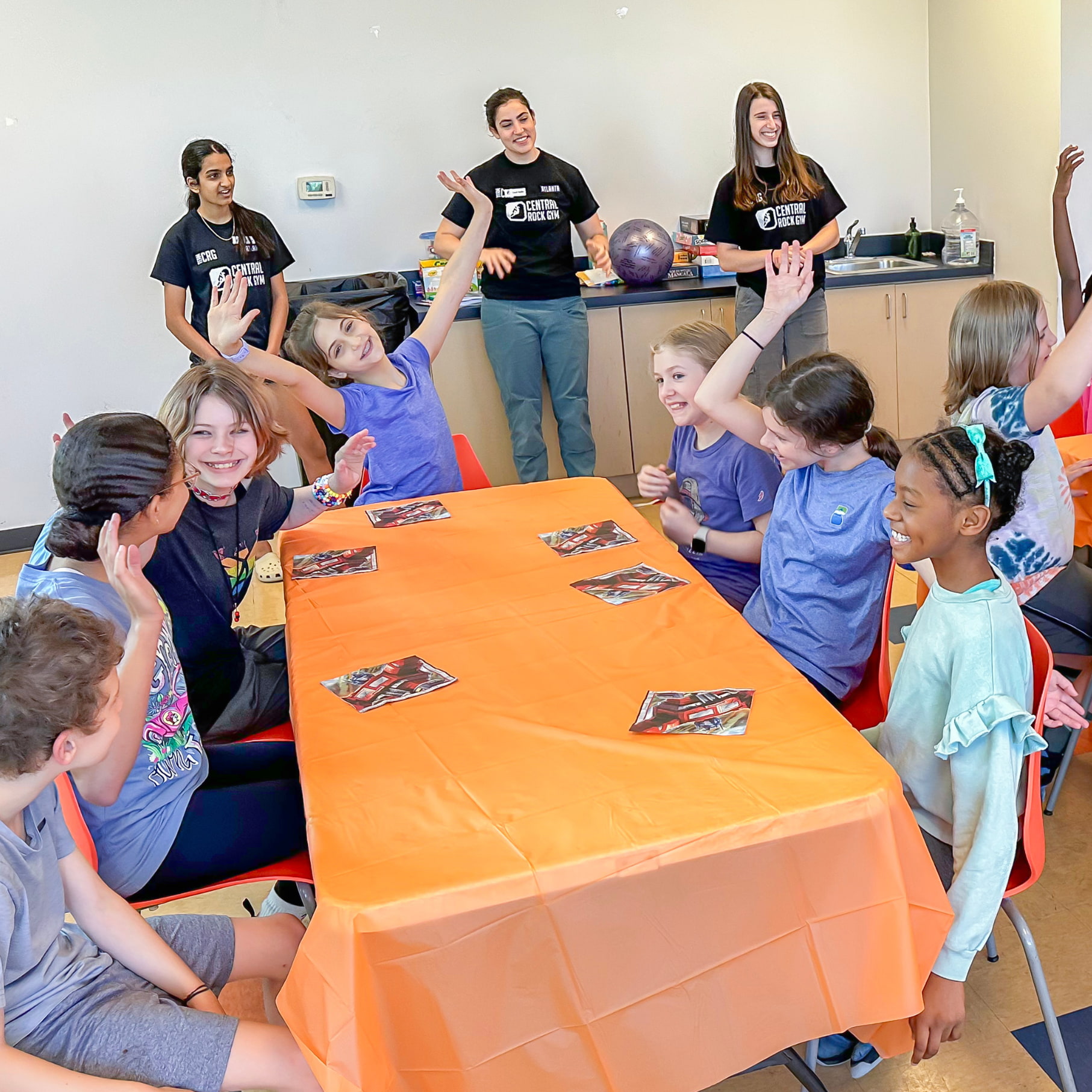 A group of young girls smile and cheer during a birthday party at Central Rock Gym. They are sitting around a table as they prepare to sing Happy Birthday.