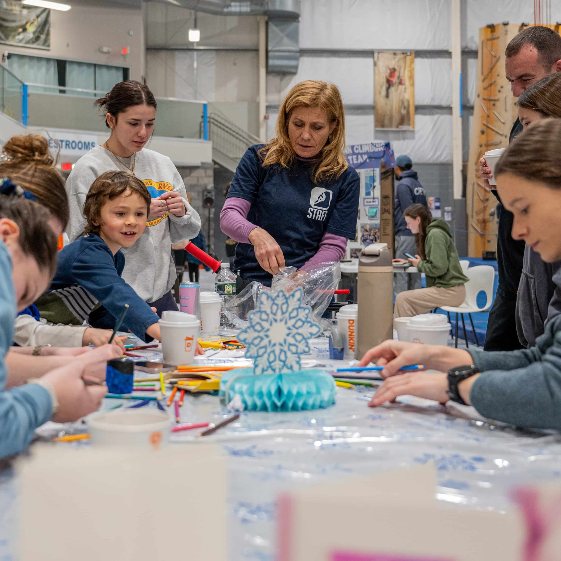 A Central Rock Gym staff member guides a group event and provides direction as everyone creates winter themed crafts at a table. The group is mixed ages all the way from young children to older adults.
