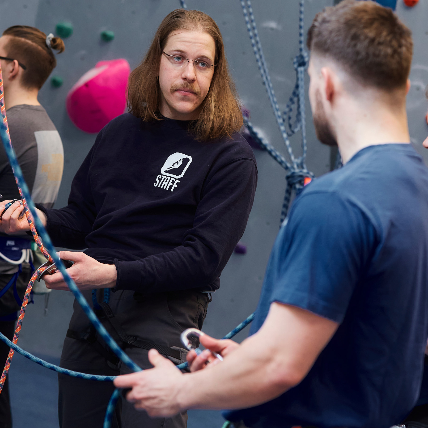 A central Rock Gym instructor (he has glasses and long hair) teaches a man with a beard how to set up a belay device during a class.