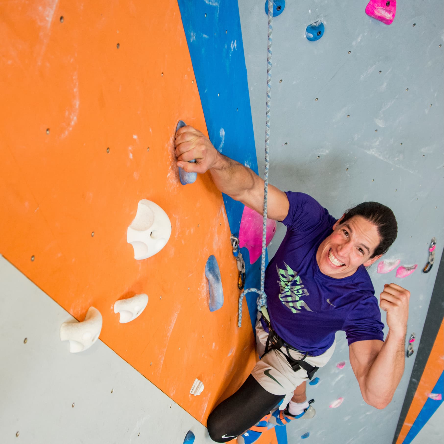 A man top rope climing while looking at camera and looking exited at central rock gym
