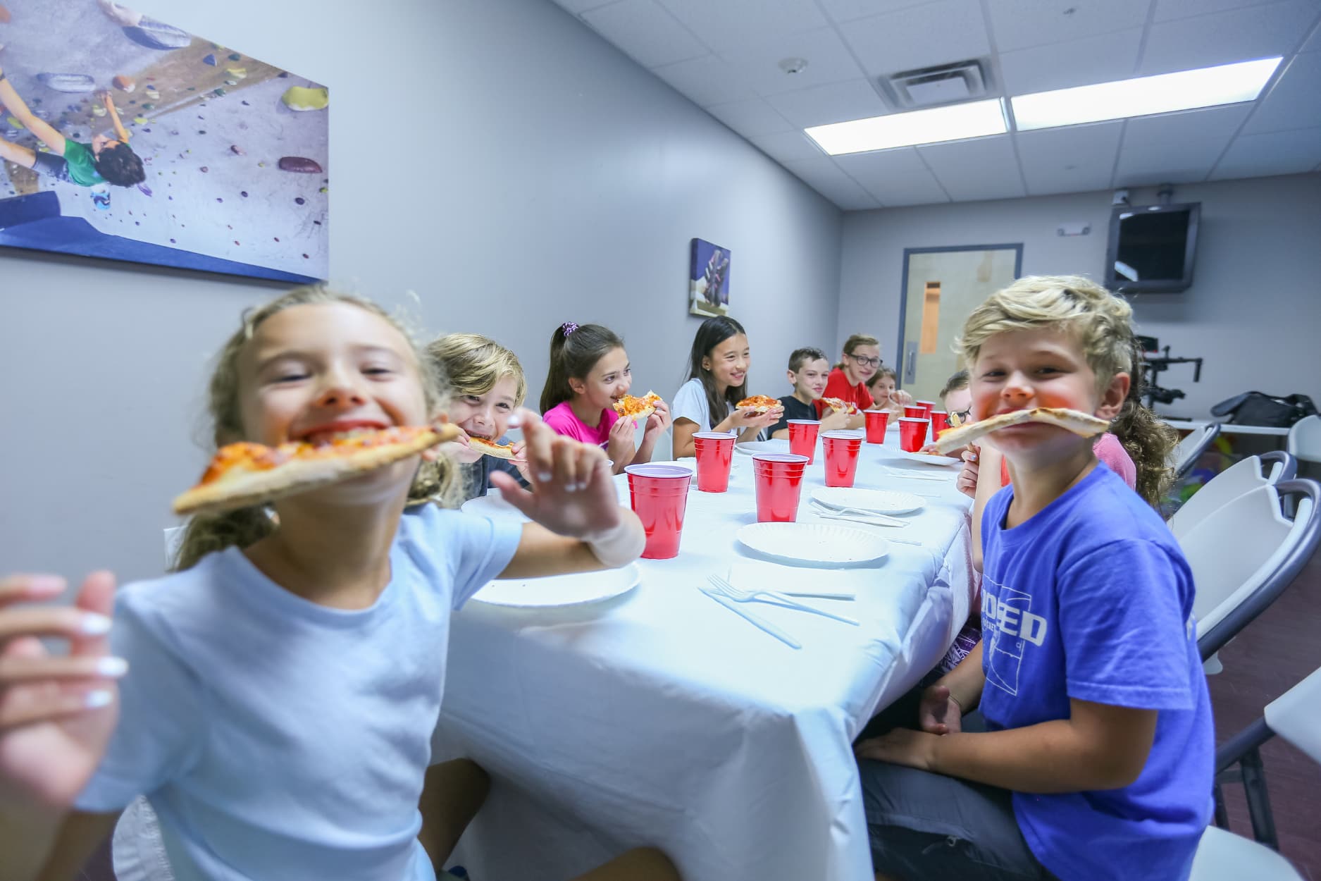 A group of kids gather around a table and eat pizza during a birthday party at central rock gym. Two kids are holding a slice of pizza in their mouths and smiling at the camera.